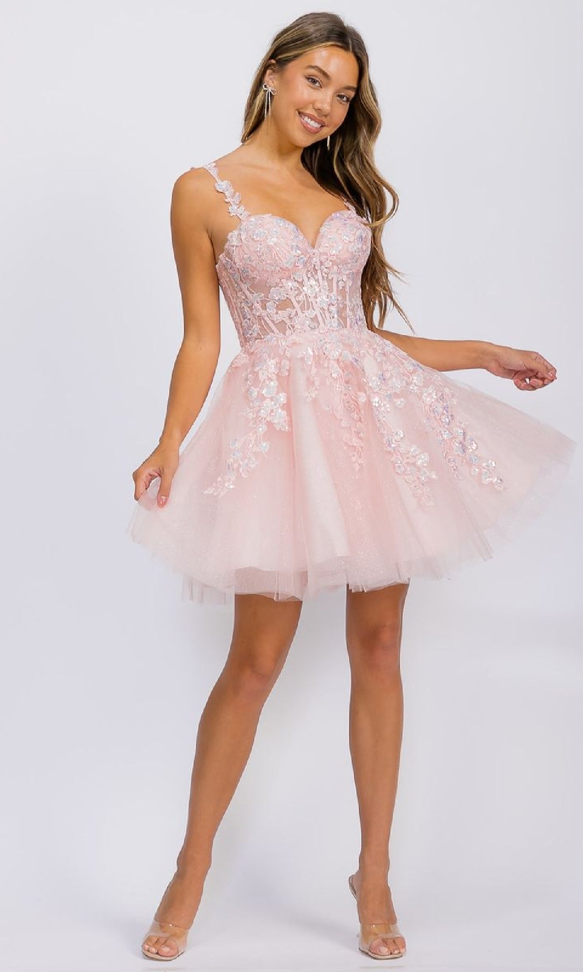 A woman wears the Eureka EK152 Short Fit-and-Flare Lace Hoco Dress, a light pink, sleeveless, knee-length design with embroidered floral lace and a tulle skirt, standing against a plain white background.